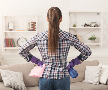 Woman with her hands on her hips, facing away from the viewer, holding cleaning equipment in a living room.